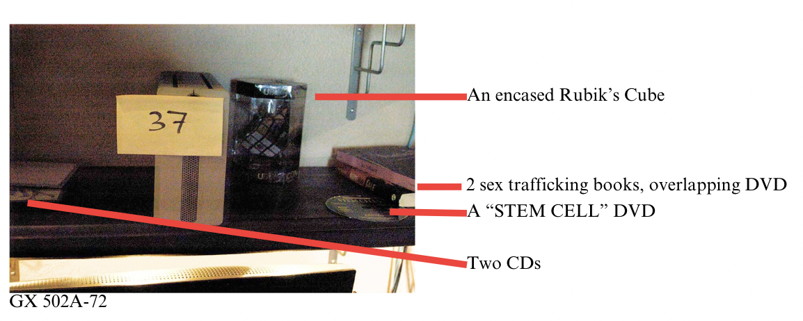 Annotated FBI search photo of the bookshelf scene with the added objects labeled — two sex-trafficking books, a STEM CELL DVD, a Rubik's cube in a case, and two CDs arranged around a single device